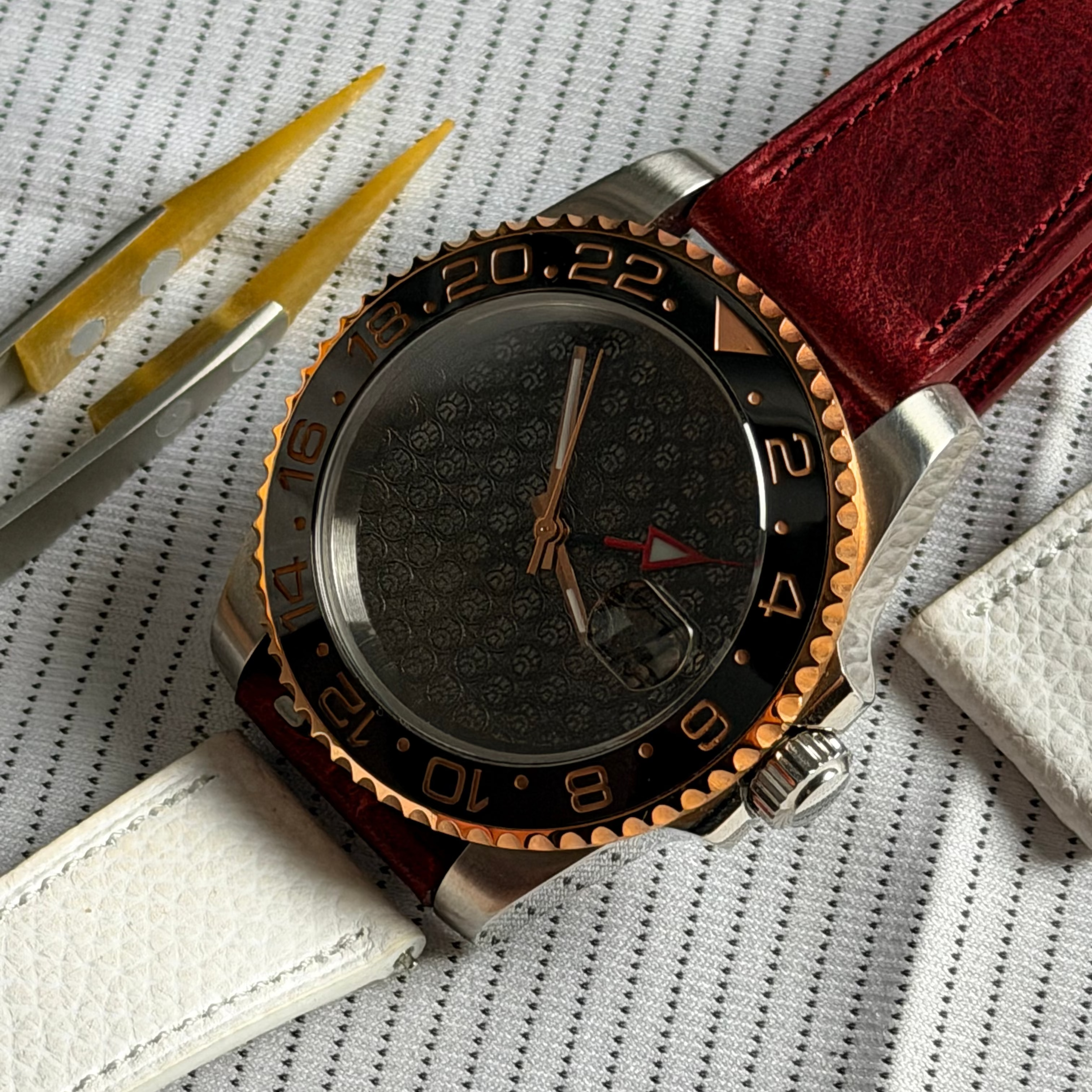 A luxury watch with a dark patterned dial, a black and rose gold bezel, and a red leather strap, displayed on a textured white background with tweezers and a white leather accessory.
