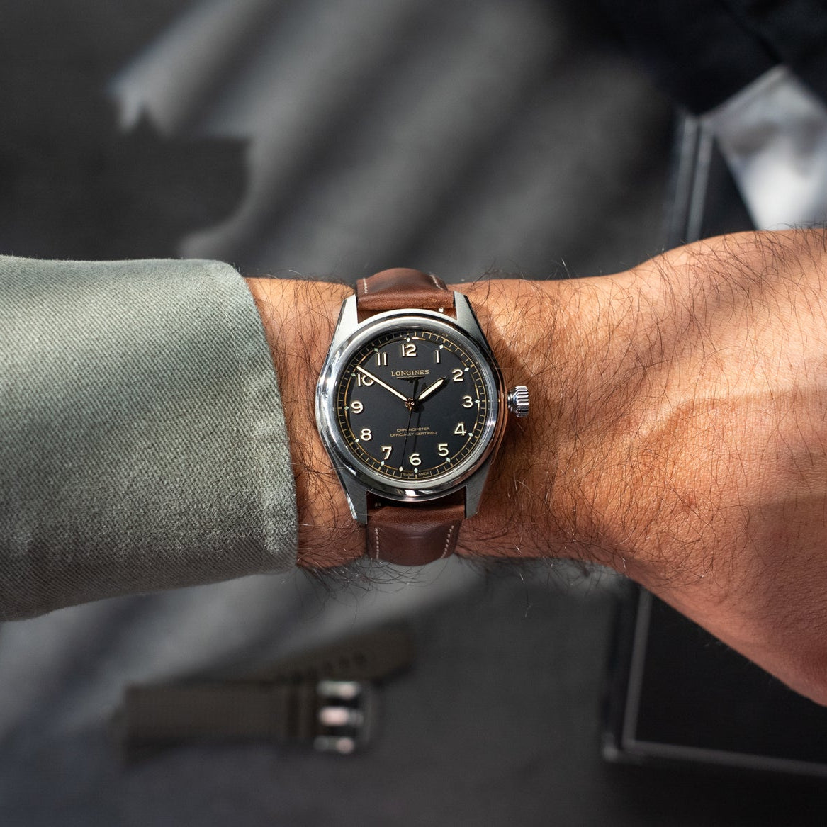 Close-up of a wrist wearing a brown leather watch with a dark face, against a blurred background.