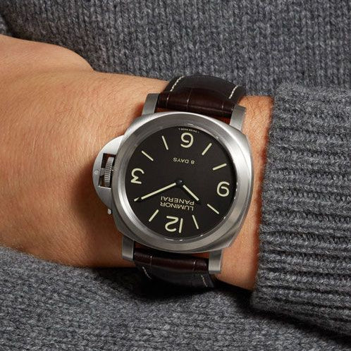 Watch with a black face and brown leather strap worn on a wrist against a gray background