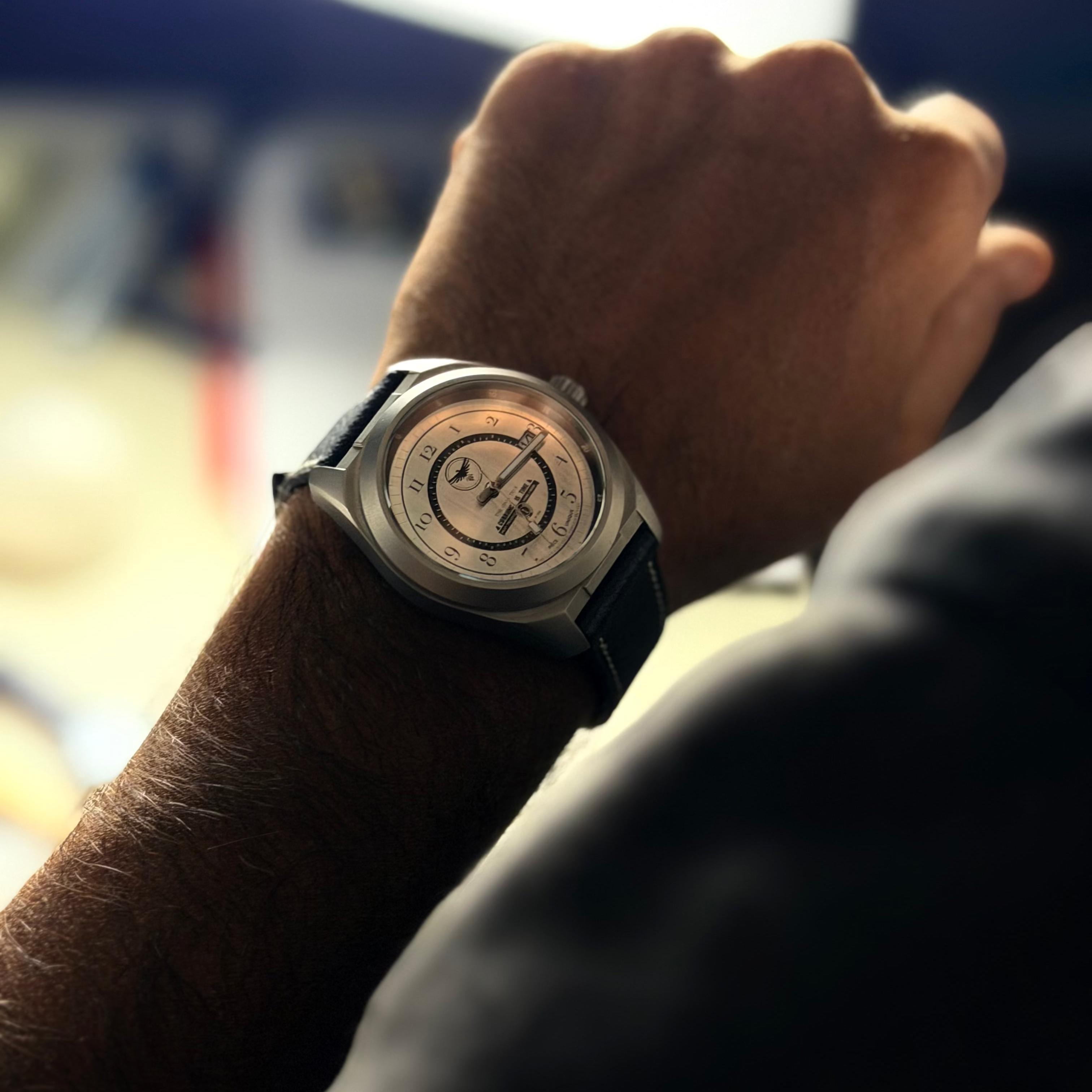A close-up lifestyle shot of a wrist wearing a silver-toned chronograph watch with a black leather strap. The watch face is open, showing the inner dial details.