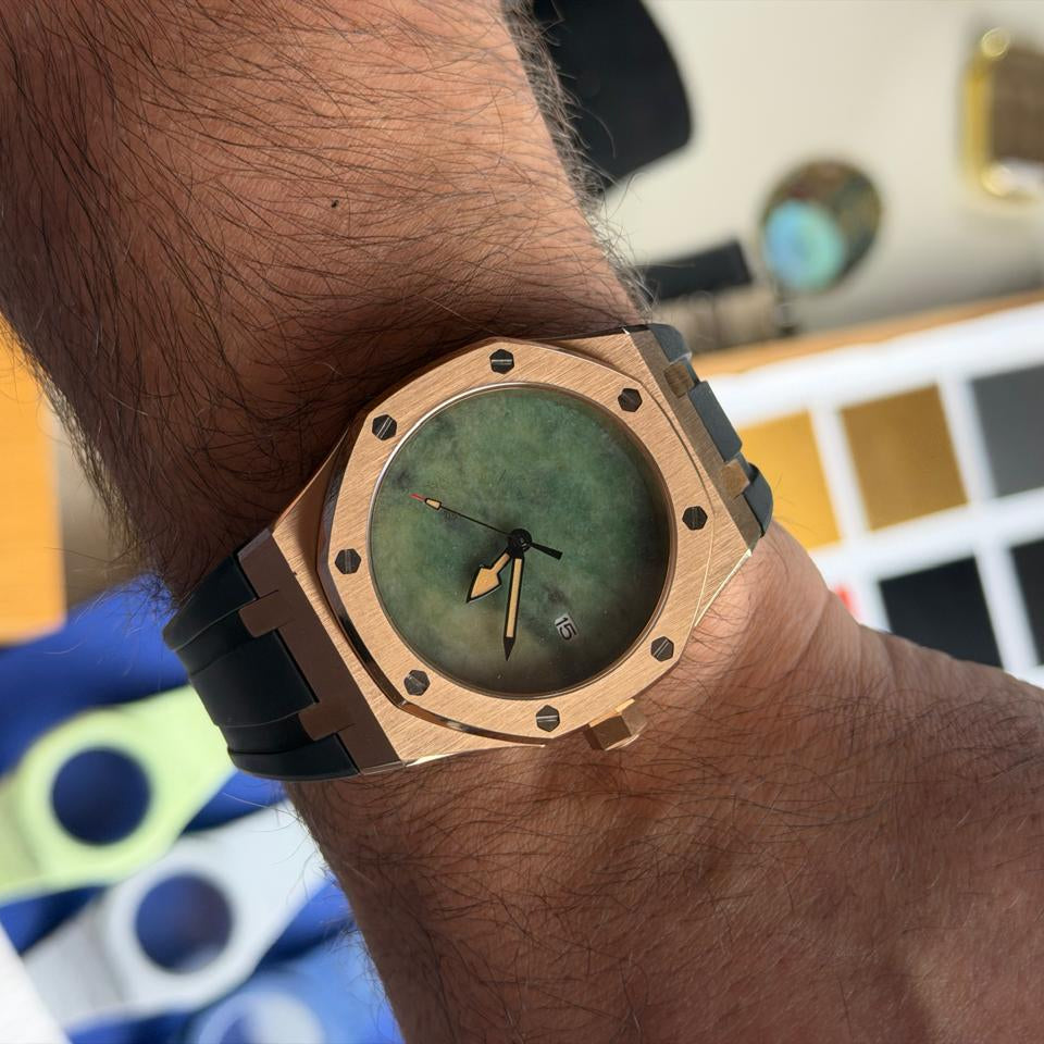 A close-up of a wrist wearing a luxury watch with a rose gold-toned octagonal case, a green dial, and a black rubber strap, set against a blurred workshop background.