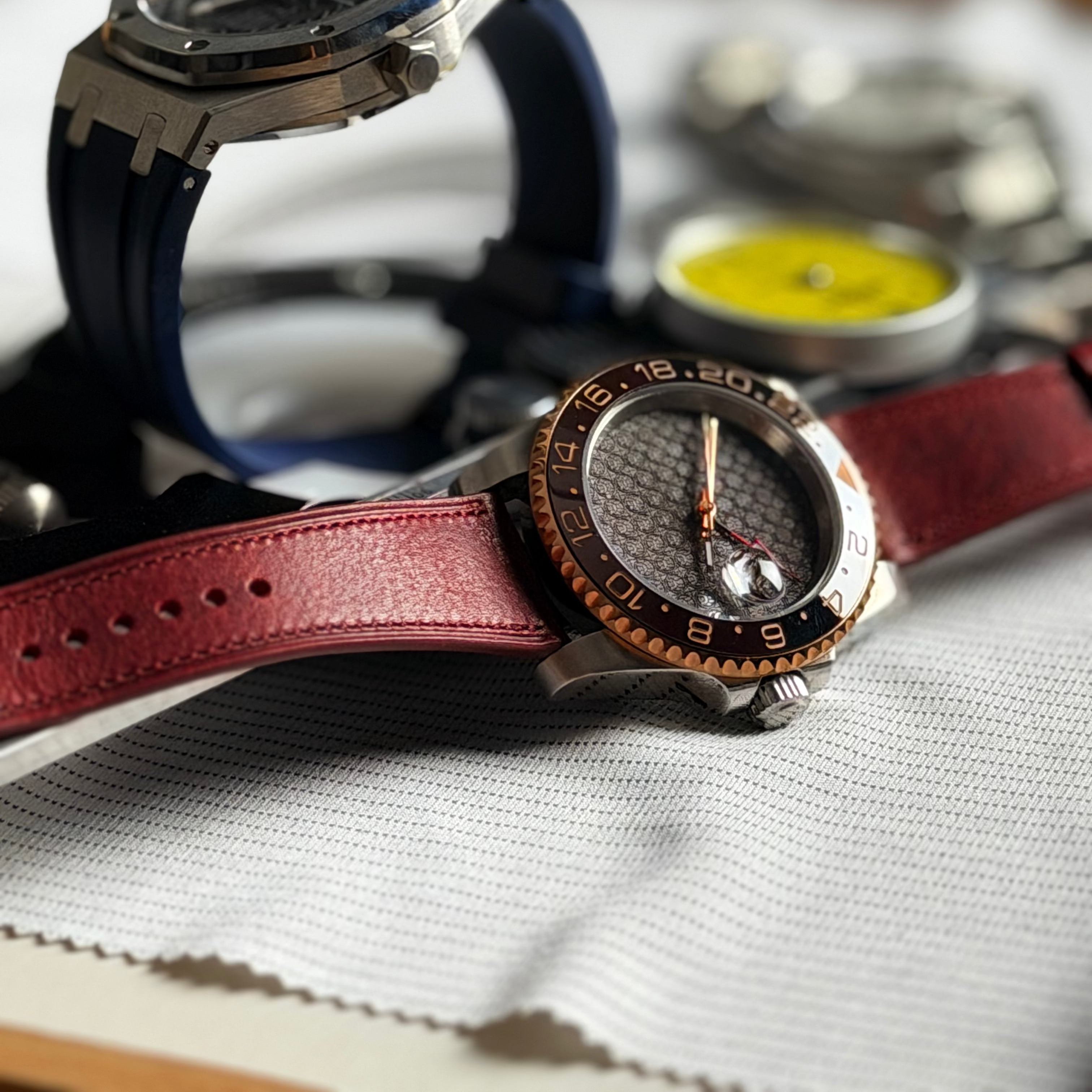 A close-up product shot of two wristwatches arranged on a white textured surface. The foreground watch has a dark dial, a bronze-toned bezel, and a burgundy leather strap.