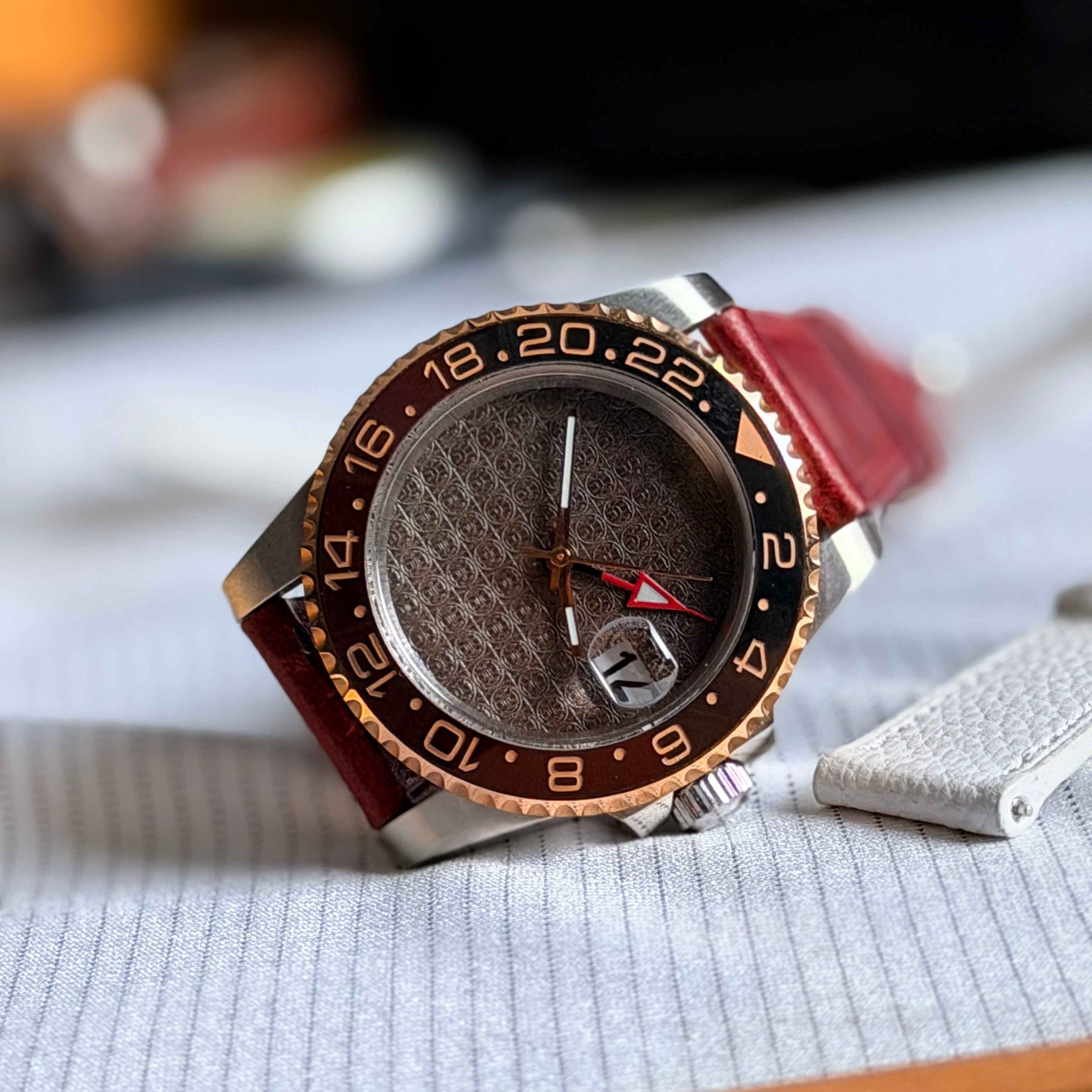 A close-up of a wristwatch with a textured brown dial, a black and rose gold tachymeter bezel, and a red leather strap, resting on a light gray textured surface.