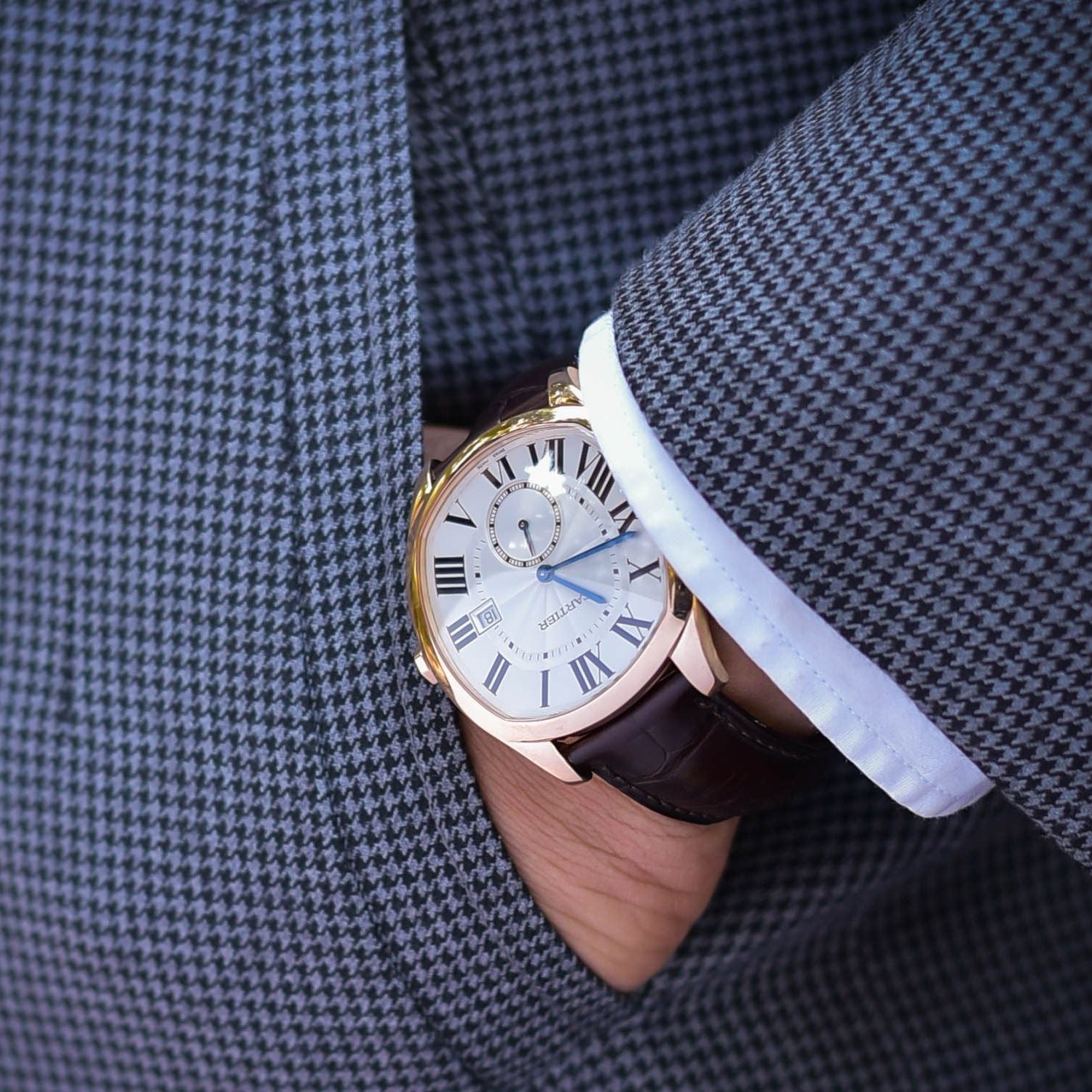 Close-up of a person wearing a watch with a detailed face and brown strap, partially hidden by a gray checkered suit.