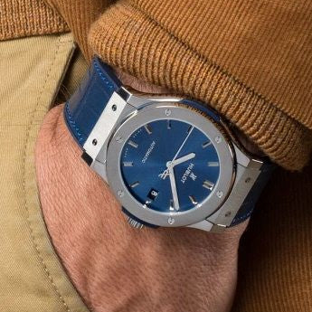 Blue watch with silver case on a wrist, wearing a brown jacket.