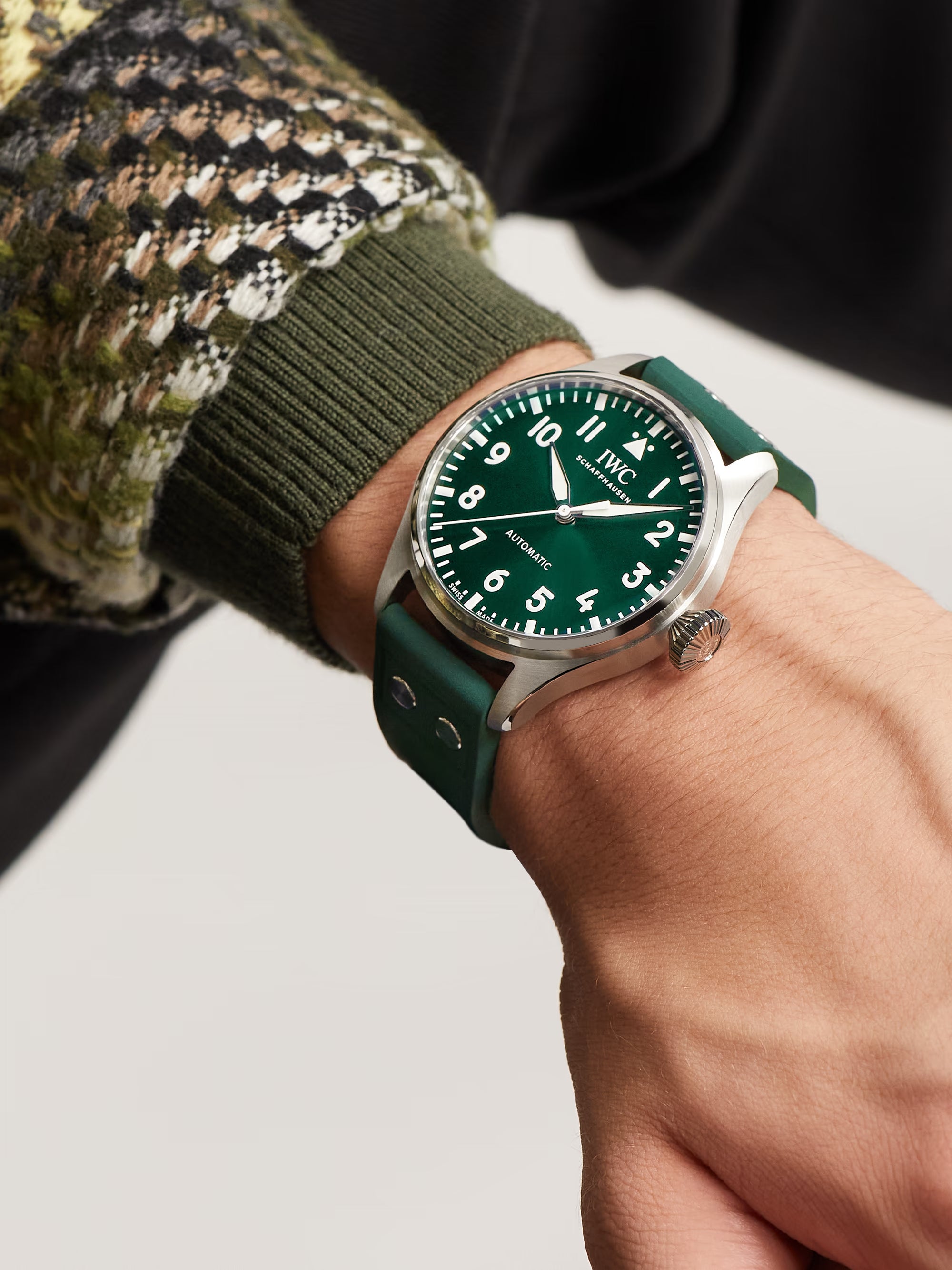 Hand wearing a green watch with a detailed face on a neutral background